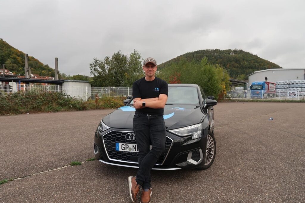 Fahrlehrer steht mit verschränkten Armen vor der Motorhaube seines Auto